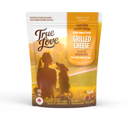 Bag of True Love dog treats with a woman smiling at her dog on the grass. Grilled Cheese flavour - golden cheddar and vine-ripened tomato.